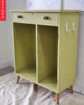 Green wooden cabinet with two open shelves, a drawer, and decorative knobs, set on tapered legs.