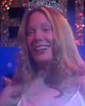 Smiling woman with a tiara on stage, glittery "Senior" sign in background.