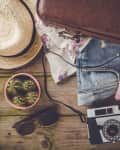 Open suitcase with summer clothes, straw hat, floral sneakers, sunglasses, camera, and small cactus on wooden floor.