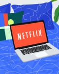 Laptop displaying Netflix on a blue bed with abstract pillow and potted plant nearby.
