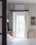 Small bedroom with a white bed, wall tapestry, and large windows with sheer curtains.