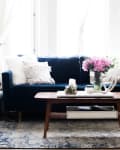 Living room with a navy sofa, pink flowers on a wooden coffee table, and a floor lamp beside stacked blankets.
