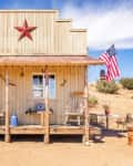 Rustic wooden cabin with a red star, rocking chairs, and an American flag in a desert setting.