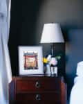 Bedside table with framed photo, lamp, and flowers next to a bed with a gray headboard and white bedding.
