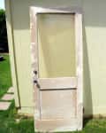 Weathered wooden door with glass panel leaning against a light yellow shed on a grassy lawn.