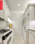 Narrow white kitchen with gas stove, tiled backsplash, and refrigerator adorned with magnets and papers.