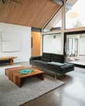 Modern living room with vaulted wood ceiling, black sofa, wooden coffee table, and large windows overlooking a patio.