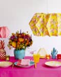 Colorful table setting with pink tablecloth, floral centerpiece, patterned plates, and origami pendant lights.
