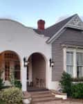 Historic house with arched porch, gray siding, and decorative trim, surrounded by greenery and outdoor lanterns.
