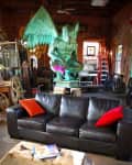 Black leather sofa with red and orange pillows in a rustic room, large green gargoyle statue, wooden coffee table.