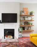 Living room with a wall-mounted TV, white fireplace, wooden shelves, yellow sofa, and patterned rug.