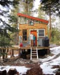 Couple standing on porch of a small wooden cabin in snowy forest, surrounded by pine trees.