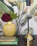 Yellow vase with red flowers, vintage books, and a gold lamp on a table against a botanical wallpaper.