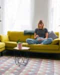 Person sitting on a yellow sofa eating, another walking by, in a living room with plants and a geometric rug.