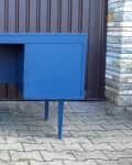 Blue mid-century modern desk with three drawers and a cabinet, set on a paved outdoor area.