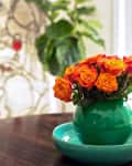 Orange roses in a green vase on a wooden table, with a leafy plant and abstract art in the background.