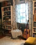 Home office with desk, computer, bookshelves, guitar, and chair by window with patterned curtains.