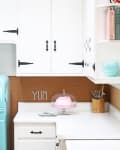 Retro kitchen with white cabinets, turquoise fridge, pink cake on stand, and "YUM" on cork backsplash.