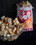 Popcorn with almonds and chocolate candies in a glass bowl next to a red and orange popcorn bag.