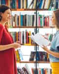 Two women talking in a library, one holding an open book, surrounded by shelves of books.