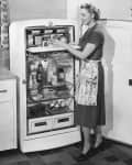 Woman in vintage kitchen placing food in an open refrigerator, wearing a dress and apron, with a bowl on the counter.