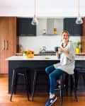Woman sitting on a kitchen stool, holding a mug, with a fruit bowl on the counter and pendant lights above.