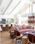 Open-concept living room with pink sofa, wooden chairs, colorful rug, and large windows overlooking greenery.