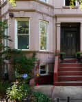 Brownstone entrance with red steps, black door, and potted plants, flanked by windows and greenery.