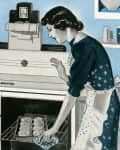 Woman in a vintage kitchen placing a tray of rolls into an oven, surrounded by blue and white decor.