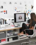 Woman working at a desk with a computer, surrounded by art and design materials on a white wall.