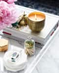 Lit candle in a gold holder on a marble tray with pink flowers, books, and a glass jar of matches.