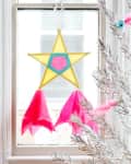 Yellow star-shaped piñata with pink and blue accents hanging in a window, next to a silver tinsel tree.