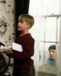 Child in red sweater holding toy near decorated Christmas tree, with a person peering through the window.