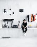 Man in black outfit sitting with a dog on a ledge in a modern room with skateboard, clothes rack, and wall art.