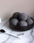 Charcoal bath bombs in a black bowl, marble soap bars, and a jar with a spoon of black powder on a marble surface.