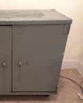 Worn gray metal cabinet with two doors and visible scratches, set on a tiled floor.
