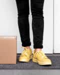 Person in yellow sneakers standing on a doormat next to a cardboard package.