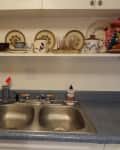 Kitchen sink with blue countertop, shelf above displaying decorative plates, mugs, and a green bottle.
