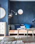 Cozy living room with a wooden daybed, blue cushions, round mirror, and hanging plants against a dark blue wall.