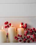 Lit white candles with red berry branches on a wooden surface.