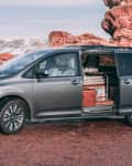 Gray minivan with sliding door open, revealing wooden interior, parked near red rock formations and snowy mountains.