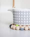 White teapot with geometric pattern and wooden handle on a beaded trivet.