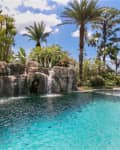 Tropical pool with cascading waterfalls, surrounded by palm trees and lush greenery under a blue sky.