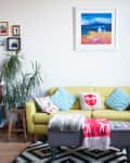 Yellow sofa with colorful cushions, surrounded by plants, art, and bookshelves in a cozy living room.