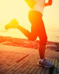 Person jogging on a paved path by the sea at sunrise, wearing athletic shoes and leggings.
