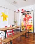 Dining area with wooden table and benches, colorful chairs, neon banana light, and vintage poster on hardwood floor.