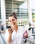 Woman in white shirt smiling at phone, holding a passport at an airport terminal.