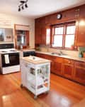 Vintage kitchen with wooden cabinets, white appliances, and a small white rolling island on hardwood floors.