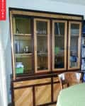 Wooden cabinet with glass doors, shelves holding books and small items, next to a round table with a green tablecloth.