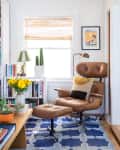 Brown leather lounge chair with ottoman on blue geometric rug, surrounded by bookshelves, yellow flowers, and a cactus.